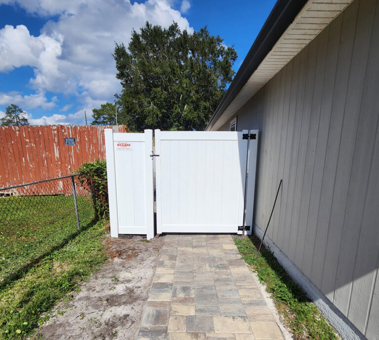 Durable white vinyl fence gate at backyard with stone path and house siding in background.