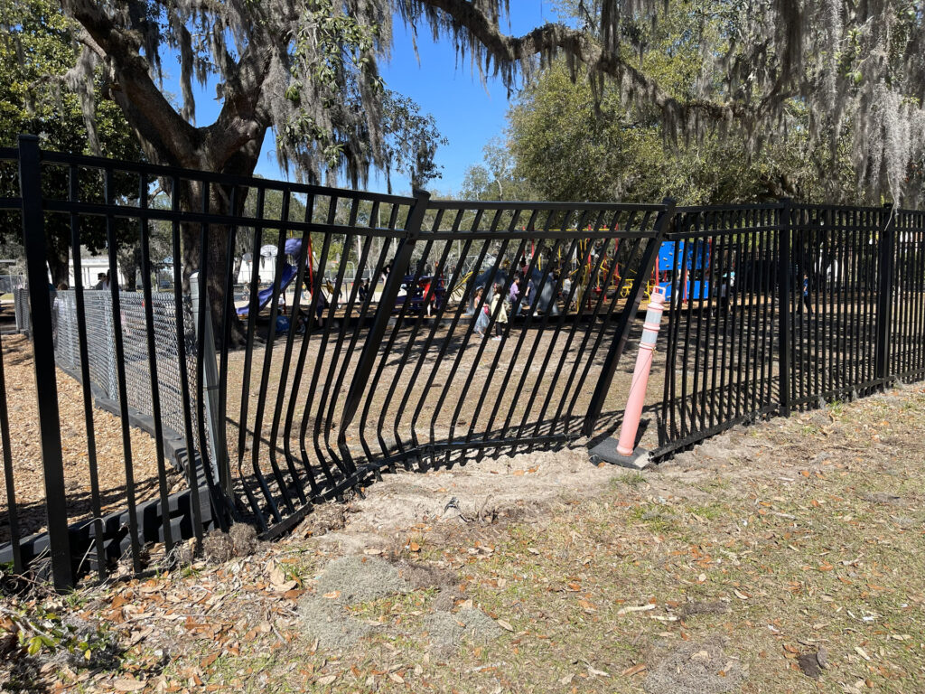 Broken black metal fence around a park with playground and trees under blue sky.
