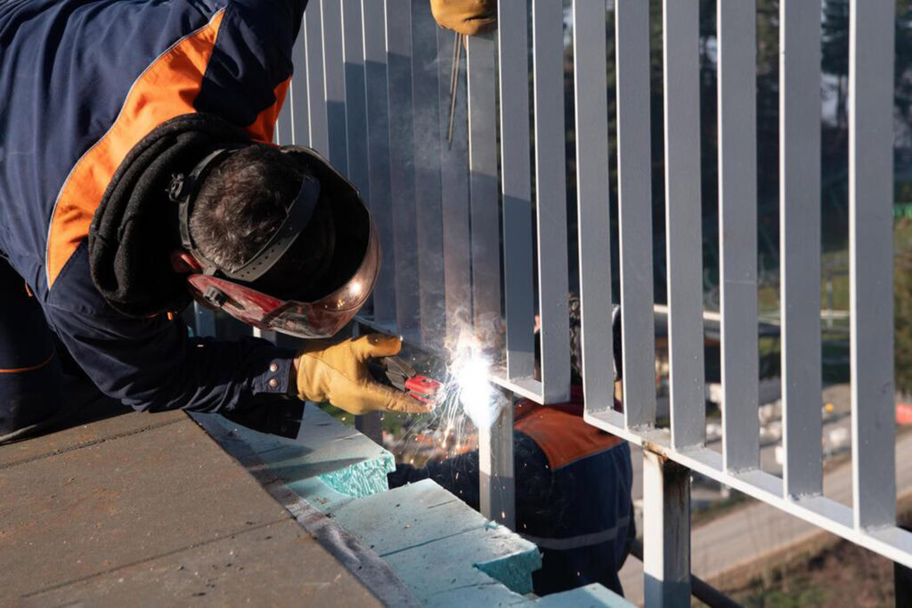 Welding worker installing a metal fence on a rooftop in bright daylight.
