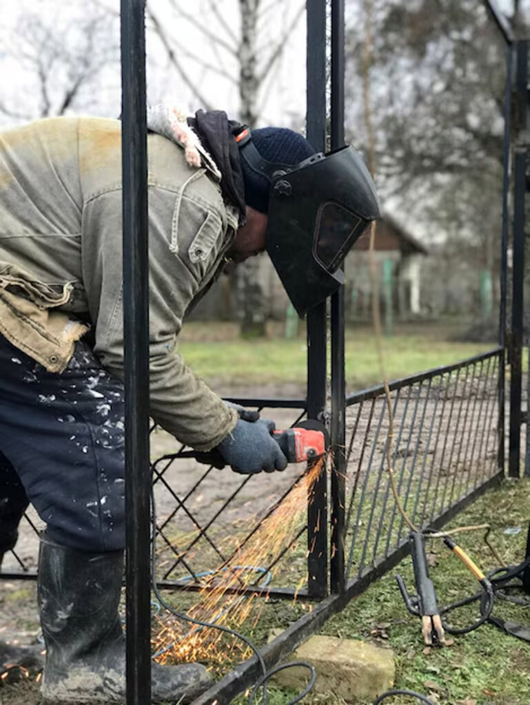 Welding metal fence with protective gear outdoors in a yard.